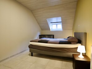 A bedroom with a sloped ceiling, a skylight, a bed with multiple pillows, a bedside table with a lamp, and a radiator. - Nice little cottage in the heart of Dinant! (Dinant)