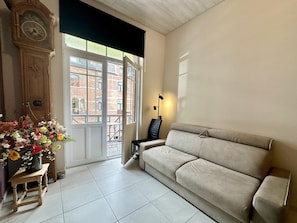 A living room with a beige sofa, a wooden side table, a clock, and a window with a view of a building. - Group house in a typical style in Dinant (Dinant)