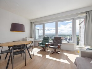 Un espace de vie moderne comprenant un coin repas, deux chaises, un canapé, une grande fenêtre donnant sur la ville et une lampe suspendue.