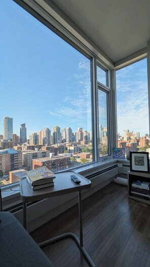 A room with a large window offering a city view, a desk with books and a remote control, and a radiator. - Entire Modern 2BR Apartment. City View. Walk to Downtown. (Calgary)