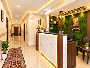 Een hotel receptie met een marmeren receptiebalie, een groene wand met planten, en een bord met de tekst 'Welcome Have a Nice Stay'.