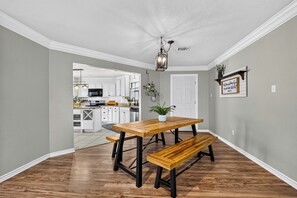 A dining area with a wooden table and bench, a kitchen in the background, and a ceiling light fixture. - Charming Spacious Retreat - 5 mins from The Woodlands Mall & i45 (Conroe)