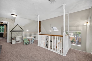 A spacious room with a high ceiling, a white staircase, a play area with a house-shaped bookshelf, and a large window offering a view of the outdoors. - Charming Spacious Retreat - 5 mins from The Woodlands Mall & i45 (Conroe)