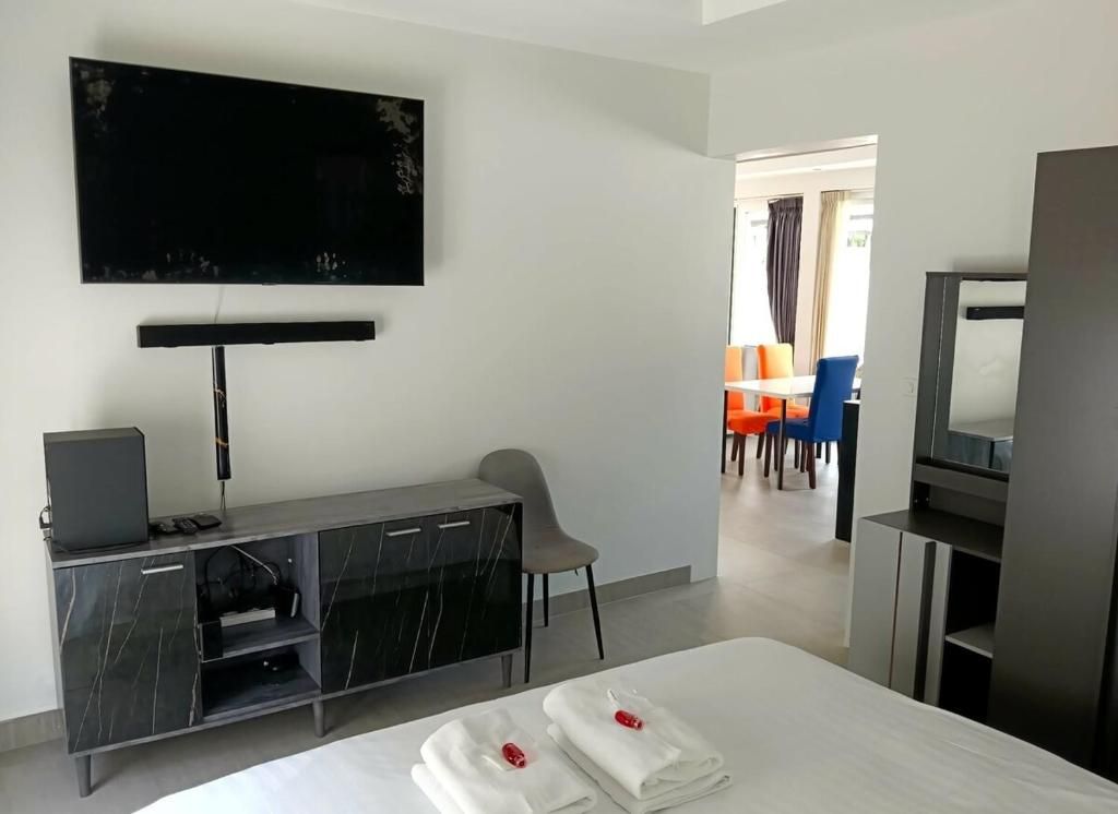 A hotel room with a bed, a TV mounted on the wall, a desk with a chair, and a dining area visible through an open door.
