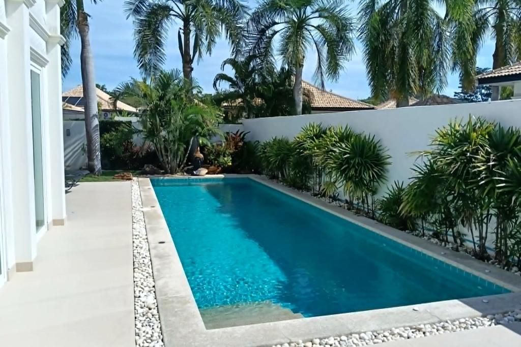 Une piscine rectangulaire à l’eau d’un bleu cristallin, entourée d’un mur blanc et bordée d’une rangée de palmiers.