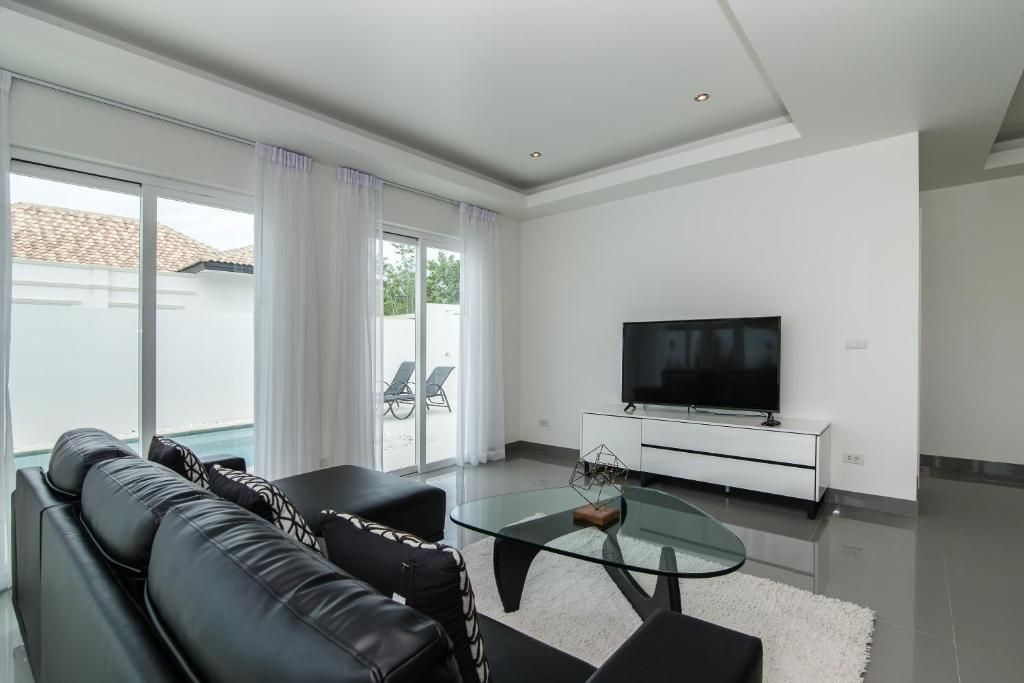Un salon moderne avec une table basse en verre, un canapé en cuir noir, un téléviseur à écran plat posé sur un meuble blanc, et une porte-fenêtre coulissante donnant sur un balcon aménagé avec du mobilier d’extérieur.