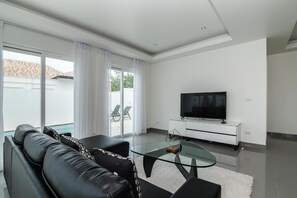 A modern living room with a glass coffee table, black leather sofa, a flat-screen TV on a white cabinet, and a sliding glass door leading to a balcony with outdoor furniture. - Orchid Paradise Homes OPV314 (Hua Hin)