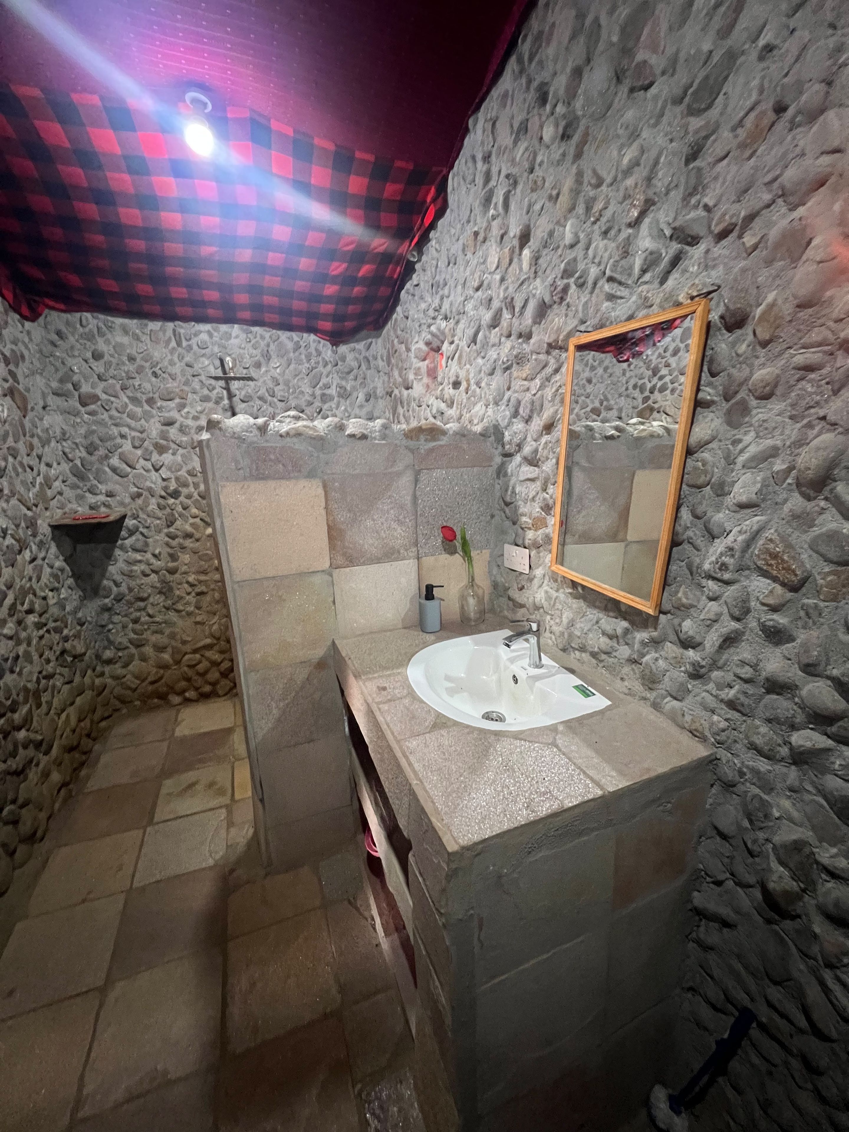 A bathroom with a stone wall, a sink, a mirror, and a red checkered cloth.