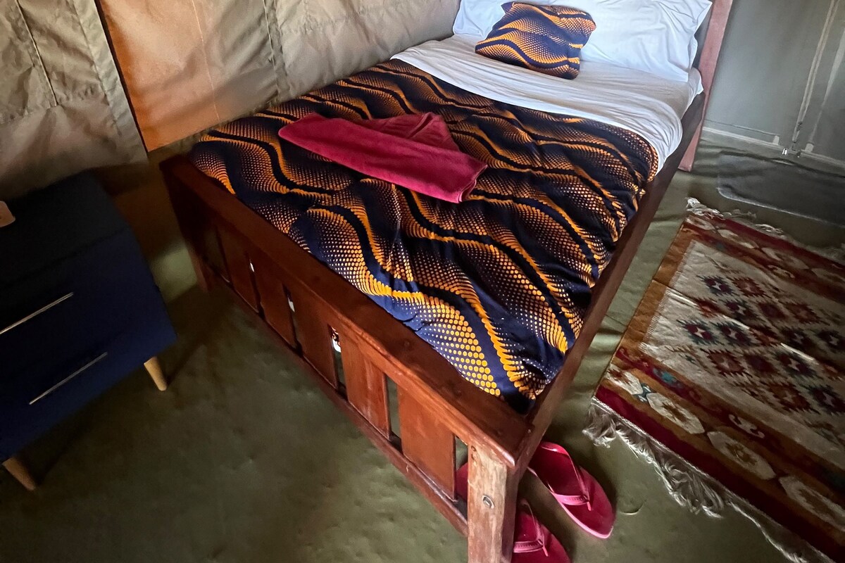 A single bed with a patterned bedspread, a wooden headboard, and a pair of pink slippers on the floor.