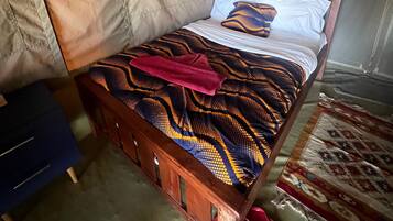 A single bed with a patterned bedspread, a wooden headboard, and a pair of pink slippers on the floor.
