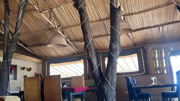 A room with a thatched roof, wooden beams, and a central tree trunk. There are tables and chairs arranged for dining.