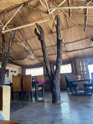 A room with a thatched roof, wooden beams, and a central tree trunk. There are tables and chairs arranged for dining. - Samara Wild Camp - Maasai Mara (Sekenani)