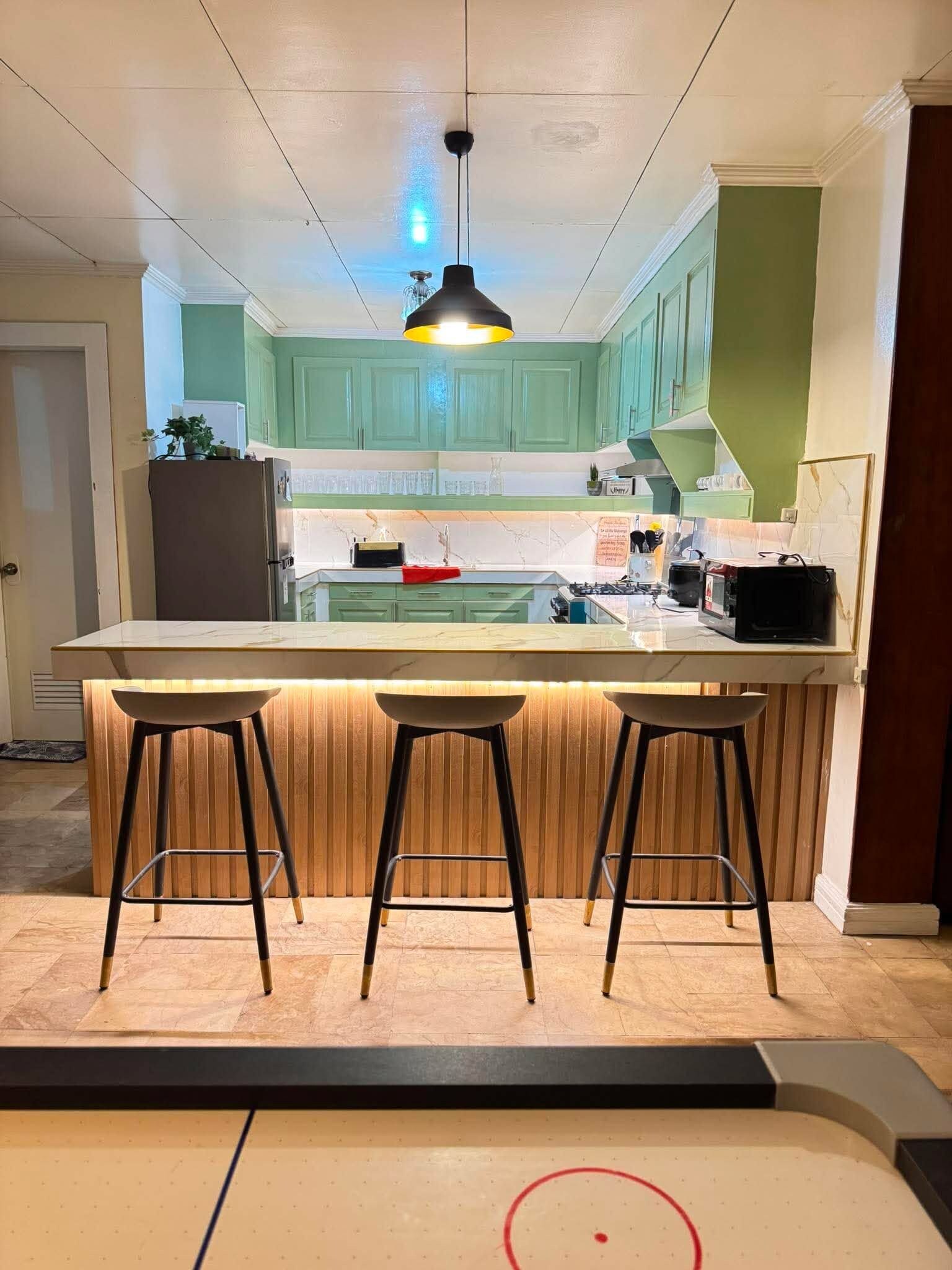 A kitchen with a central island, bar stools, a pendant light, and green cabinets.