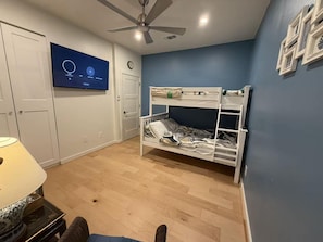 Habitació amb lliteres, ventilador de sostre, televisió penjada a la paret i làmpada sobre una taula.