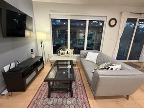 Een moderne woonkamer met een grijze bank, een glazen salontafel, een flatscreen televisie aan de muur en een groot raam met uitzicht.