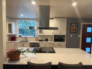 A modern kitchen with white cabinets, a stainless steel range hood, a microwave, and a large window providing a view of trees.