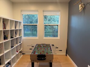 A room with a foosball table, a bookshelf, and two windows with a view of trees.