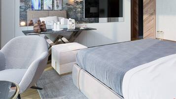 A modern hotel room with a large bed, a desk with a computer and lamp, a chair, and artwork on the wall.