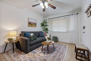 Ein Wohnzimmer mit grauem Sofa, einem kleinen runden Tisch, einer Stehlampe, einem Bild an der Wand, einem Fenster mit Jalousien und einem Deckenventilator.