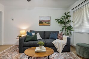 Ein Wohnzimmer mit grauem Sofa, einem runden Holztisch, einem Blumentopf und einem Bild an der Wand.