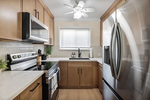 A compact kitchen with a built-in oven, a stainless steel refrigerator, wooden cabinets, and a window with blinds. - Cozy and Modern 1 Bed 1 bath Travelers Dream Home in Downtown Mountain View (Mountain View)