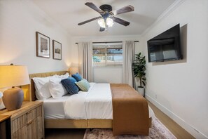 Une chambre à coucher comprenant un lit, une table de chevet, une lampe, un ventilateur de plafond, une fenêtre avec des stores, un téléviseur fixé au mur et une plante en pot.