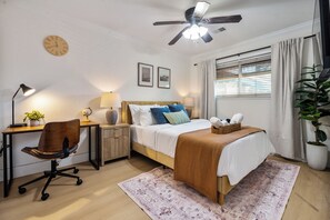Ein Hotelzimmer mit Bett, Schreibtisch, Stuhl, Lampe, Deckenventilator und einem Fenster mit Vorhängen.