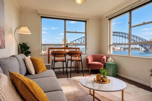 A modern living room with a grey sofa, two armchairs, a coffee table, and large windows offering a view of a bridge and cityscape. - 1BR Harbour View Nest Opal (McMahons Point)