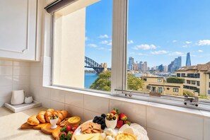 A kitchen with a view of a cityscape, including a bridge and buildings, and a variety of food items on the counter. - 1BR Harbour View Nest Opal (McMahons Point)