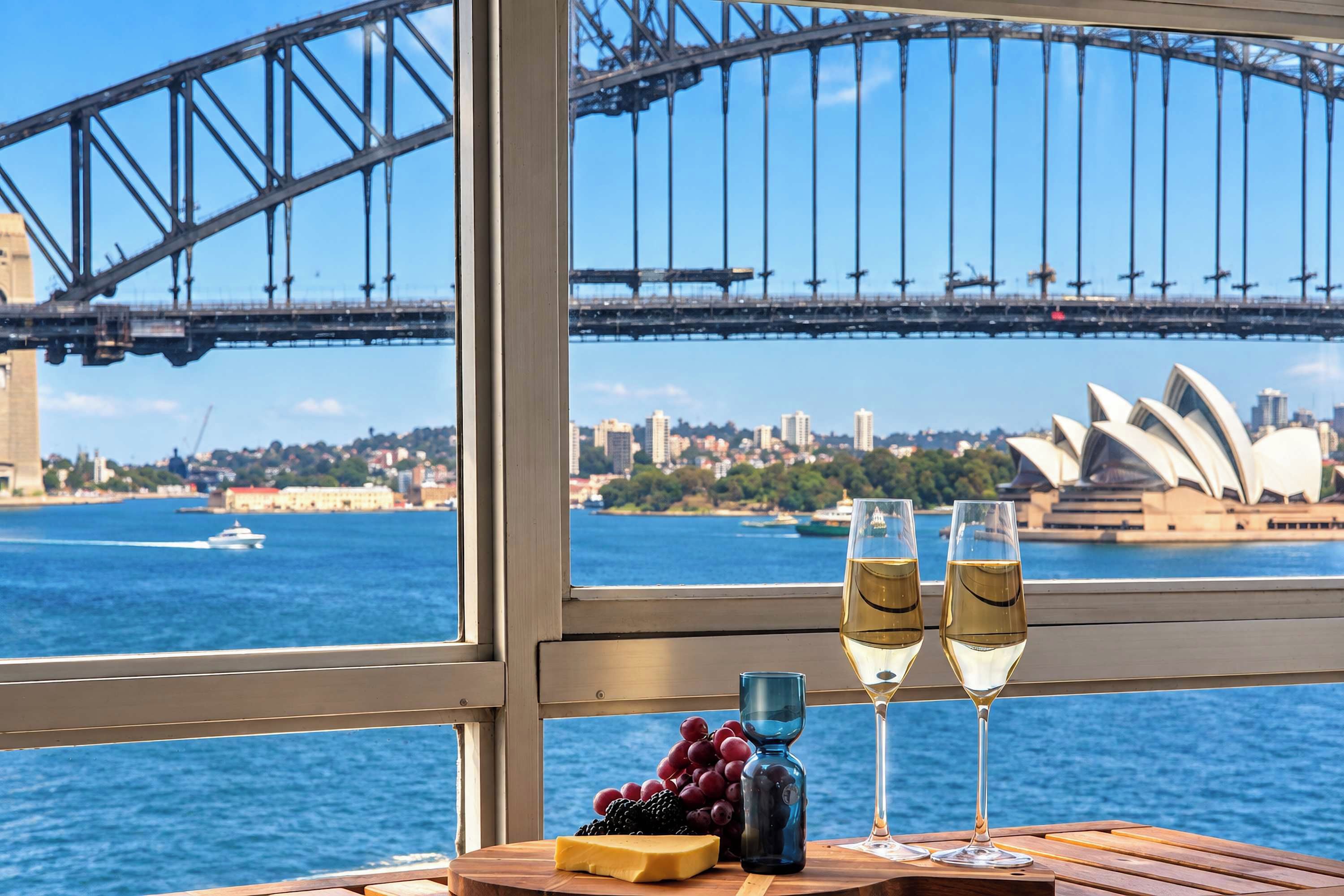 A table with a bottle of wine, two glasses, and a bowl of grapes, with a view of the Sydney Opera House and Sydney Harbour Bridge.