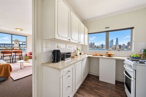A modern kitchen with white cabinetry, a gas stove, and a view of the city skyline through a large window. - 1BR Harbour View Nest Opal (McMahons Point)