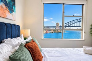 A bedroom with a bed, pillows, a nightstand, a lamp, and a window with a view of a bridge and water. - 1BR Harbour View Nest Opal (McMahons Point)