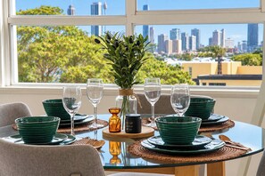 A dining table set with green plates, clear glasses, and a vase with greenery, overlooking a cityscape. - 1BR Harbour View Nest Opal (McMahons Point)