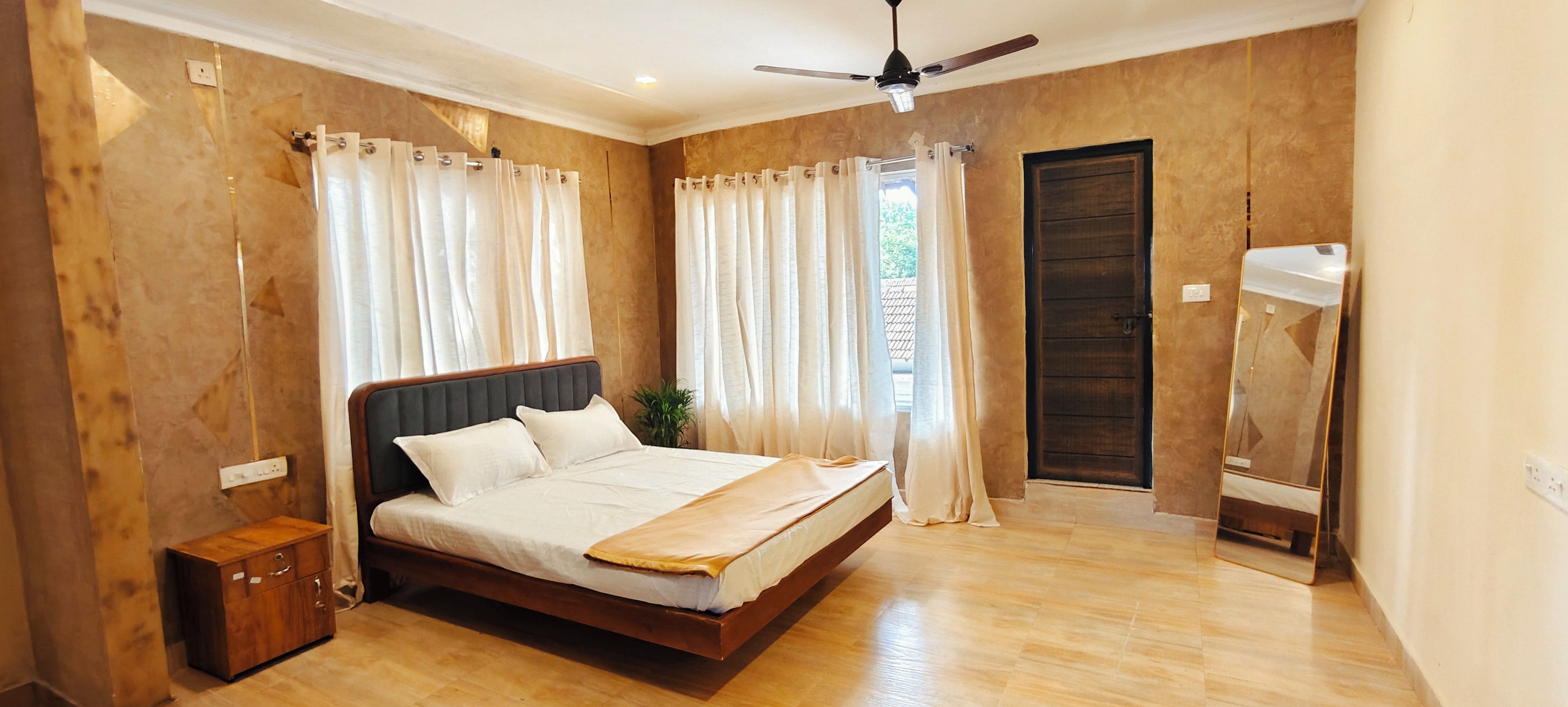 Une chambre à coucher avec un lit en bois, des rideaux blancs, un ventilateur de plafond et une porte en bois.