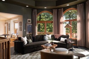 A cozy living room with a dark-colored sofa, a coffee table, and a round ottoman. There are large windows with curtains, a wooden deck visible through the windows, and a staircase leading to an upper level. - Luxe Cabin •Sauna•Hot Tub•Theater•Arcade•6 Kings (Galena)