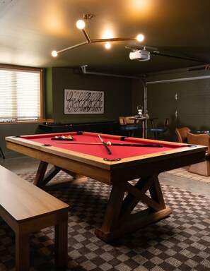 A pool table with a red felt surface, a billiards cue, and balls. There is a projector mounted on the ceiling, and a framed artwork on the wall. - Luxe Cabin •Sauna•Hot Tub•Theater•Arcade•6 Kings (Galena)