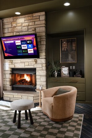 Ein Wohnzimmer mit Kamin, einem darüber montierten Fernseher, einem bequemen Sessel und einem karierten Teppich.