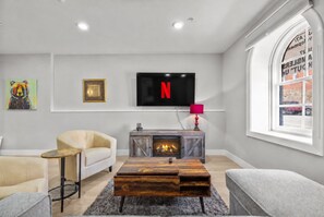 A modern living room with a fireplace, a television mounted on the wall, and a window with a view of the street.