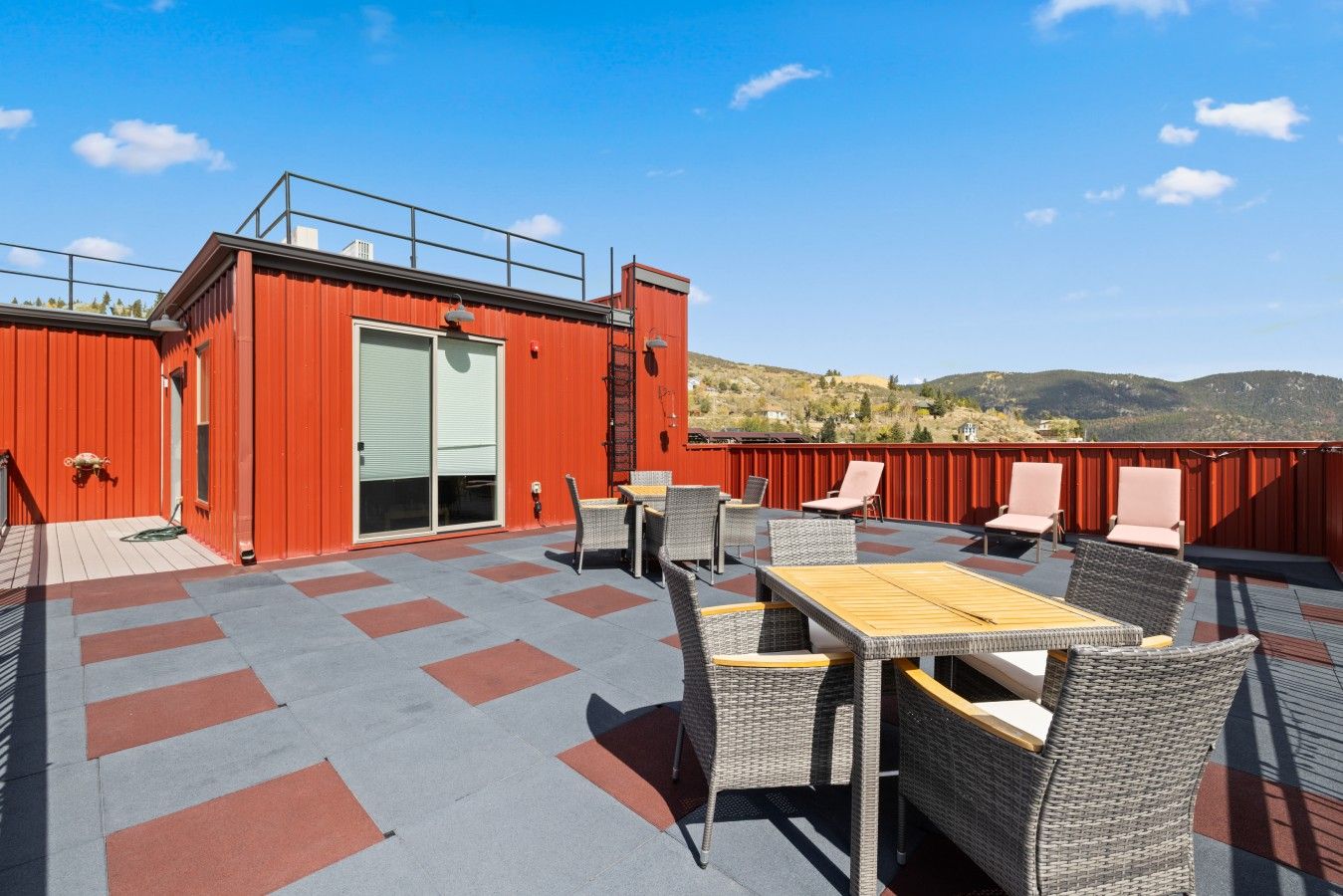 Een terras op het dak met buitenmeubilair, een rood gebouw en uitzicht op de heuvels.