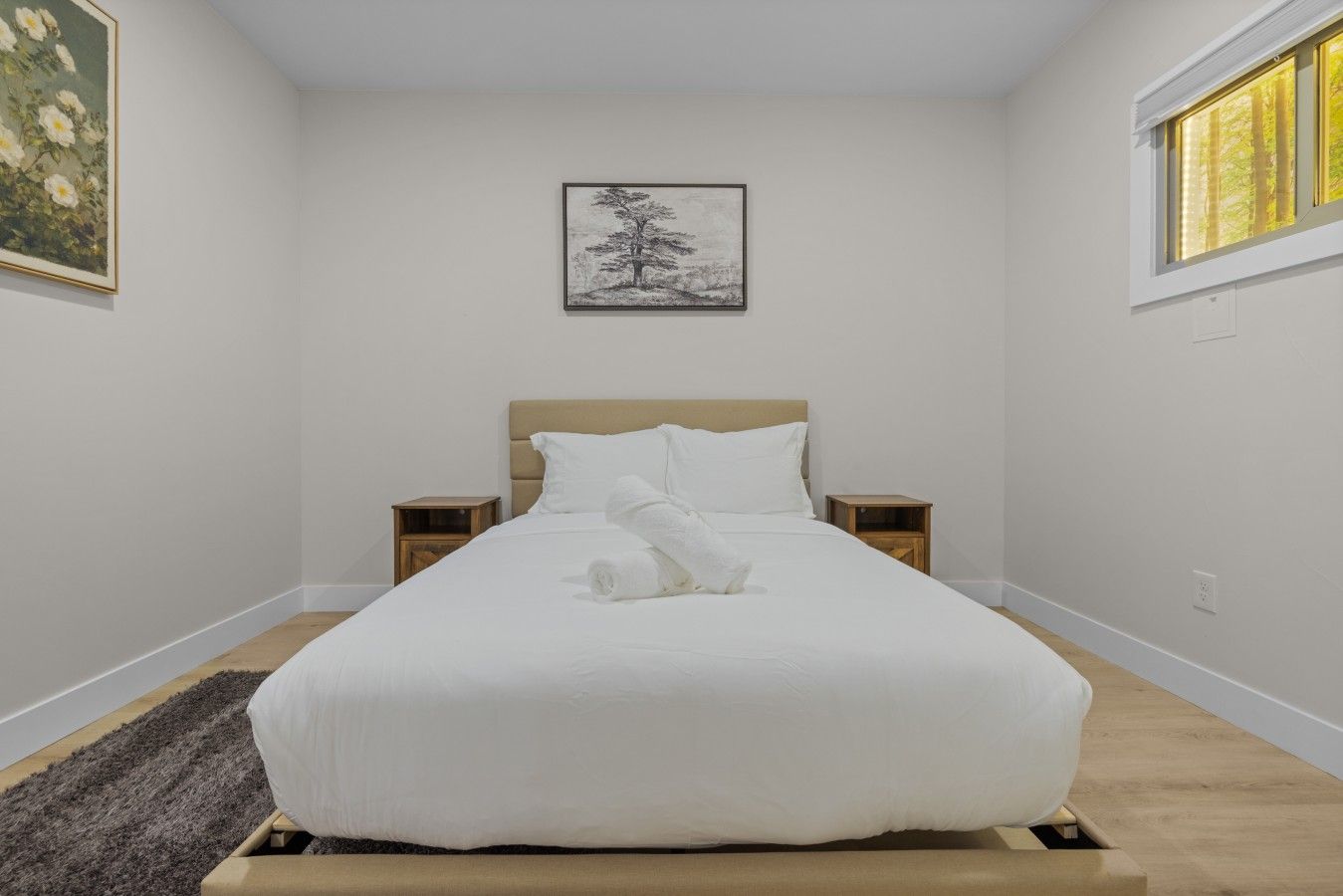 A neatly made bed with white linens and a rolled towel, two bedside tables, a framed picture on the wall, and a window with daylight.