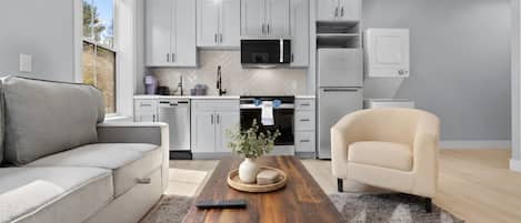 A modern kitchen with white cabinetry, a microwave, and a refrigerator. There is a wooden coffee table with a vase and a remote control.