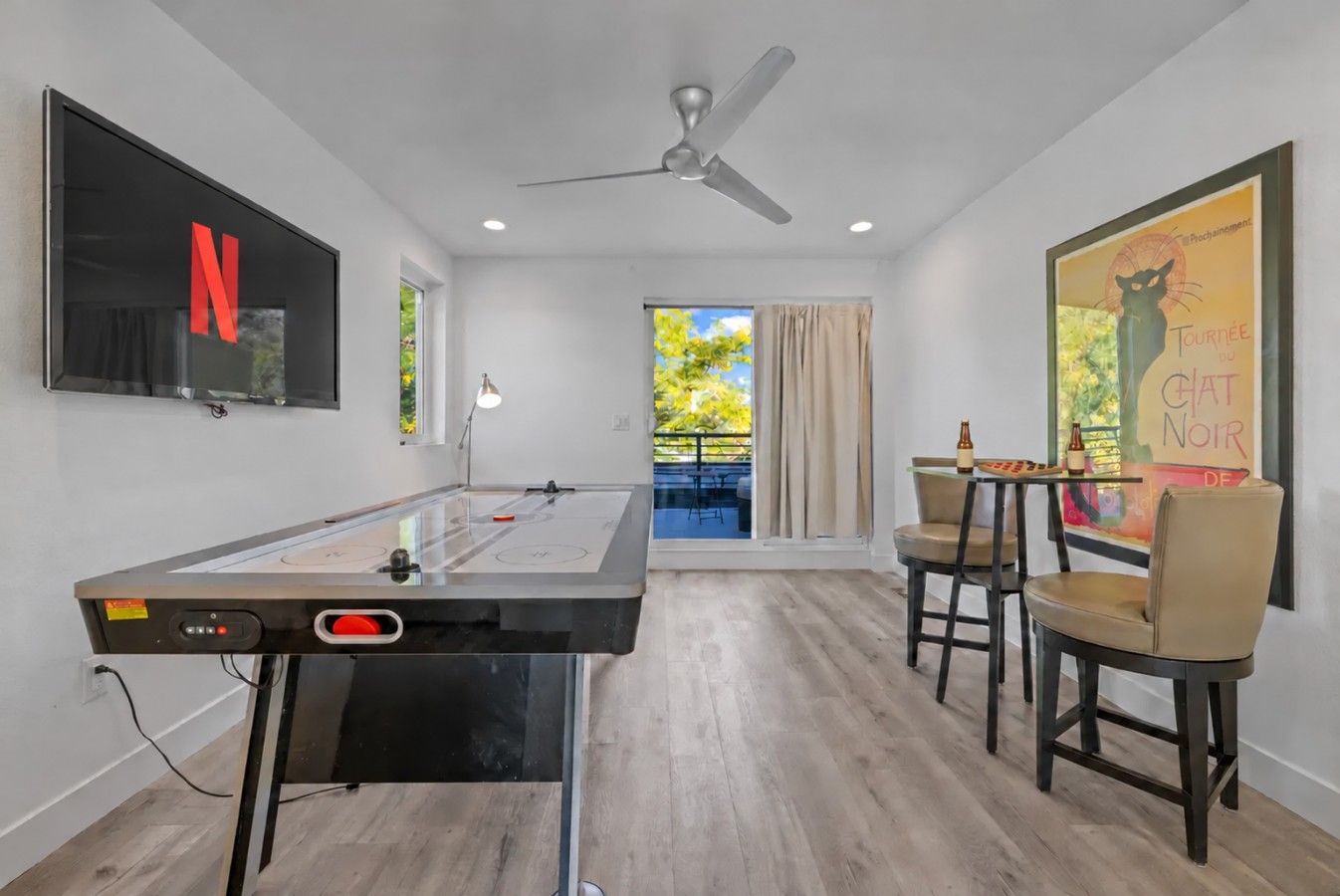 A room with a foosball table, a TV mounted on the wall, a ceiling fan, and a view of the outdoors.