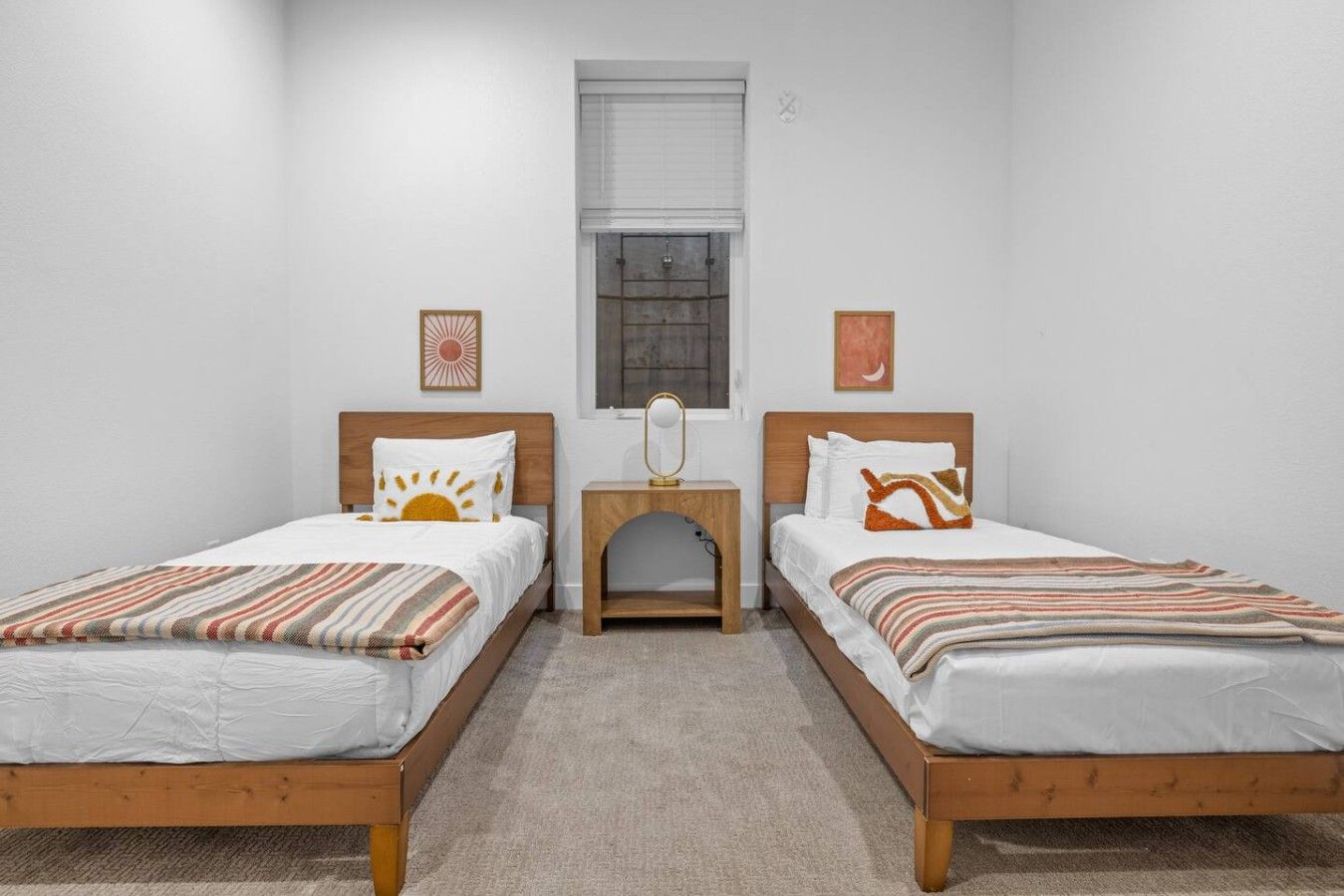A room with two wooden beds, a small wooden nightstand, a window with blinds, and two wall-mounted artworks.