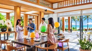 A spacious indoor area with a wooden ceiling and large windows, a dining table with chairs, and a view of the beach outside.