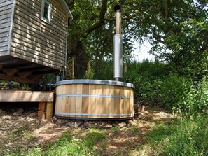 Un bain à remous en bois, doté d’une cheminée en métal, installé à l’extérieur, avec une terrasse en bois et une maison en arrière-plan.