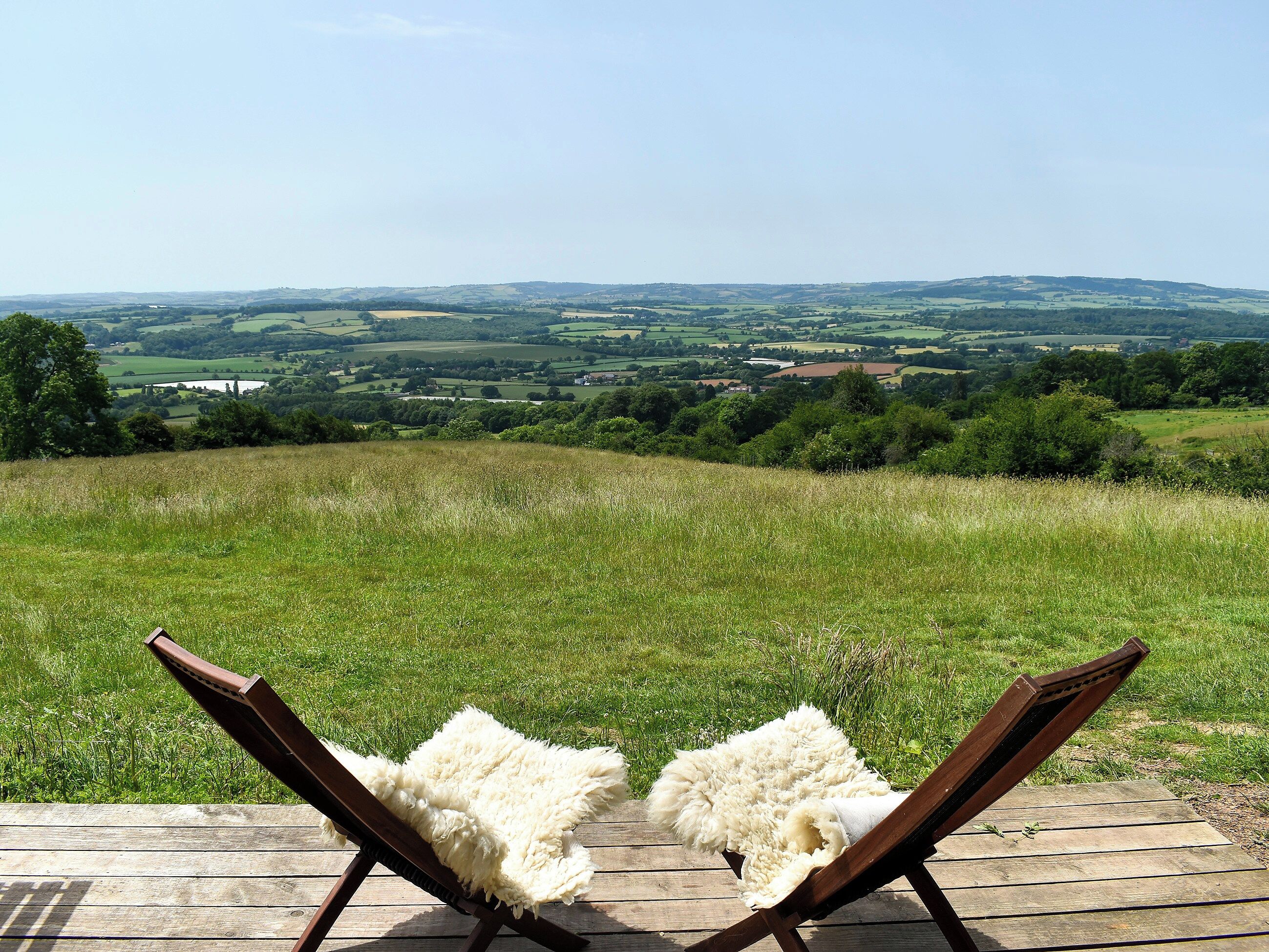 A wooden deck with two lounge chairs facing a grassy field and distant hills.
