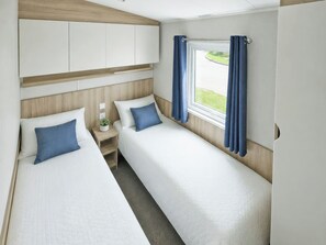 A bedroom with two beds, a nightstand, a window with curtains, and a wardrobe. - Foreshore 1 at Seashore Holiday Park (Caister-On-Sea)