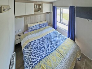 Une chambre à coucher comprenant un lit, une table de chevet, une étagère avec des bougies, une fenêtre avec des rideaux et un téléviseur fixé au mur.