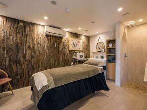 Ein modernes Schlafzimmer mit einer Holzvertäfelung, einem Bett mit dunkler Bettdecke, einem kleinen runden Tisch mit Stuhl und einem Regal mit Pflegeprodukten.