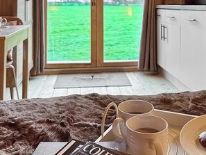 Ein gemütliches Zimmer mit Holzboden, einem Fellteppich, einem Esstisch mit einer Zeitschrift, zwei Tassen und Blick auf ein grünes Feld durch ein Fenster.